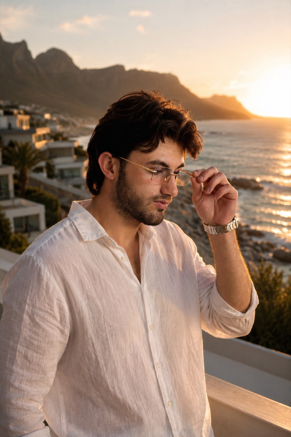 Man in a white linen shirt wearing noframe rimless sunglasses at sunset over Camps Bay